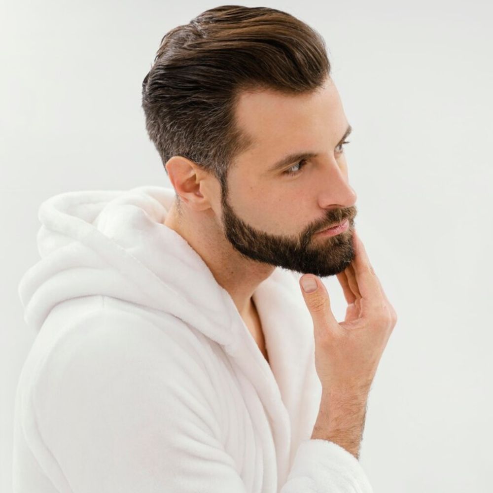 A close-up of a man's neck and jawline while using a black electric trimmer on his beard, with a light gray background.
