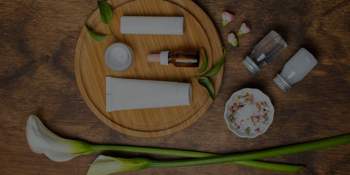 A styled flat-lay of cosmetic and skincare packaging items on a dark wood surface, including white tubes, a small amber dropper bottle, clear and frosted bottles, a small dish with dried flowers, and green leaves. A round wooden tray holds the main grouped items.