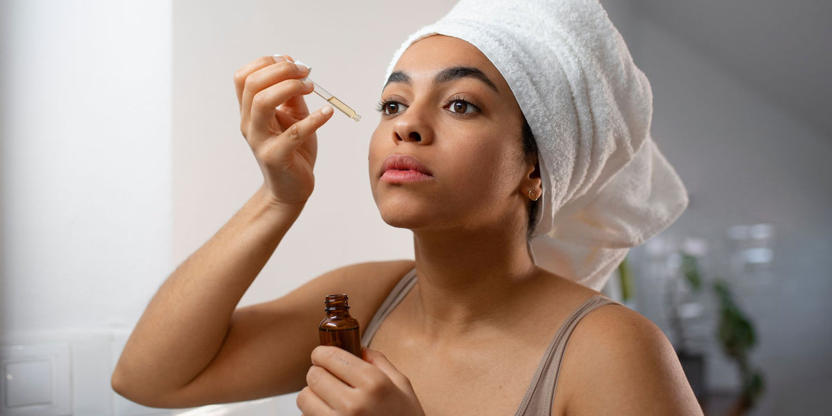 A woman with a white towel wrapped around her head applies a drop of serum from a glass dropper bottle to her eye area. She is holding an amber glass bottle in her other hand, set against a softly blurred bathroom background.