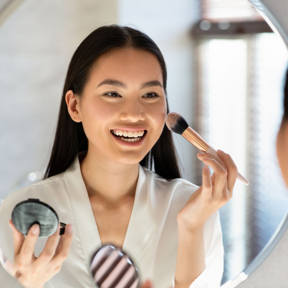 Woman applying makeup with a brush in front of a mirror